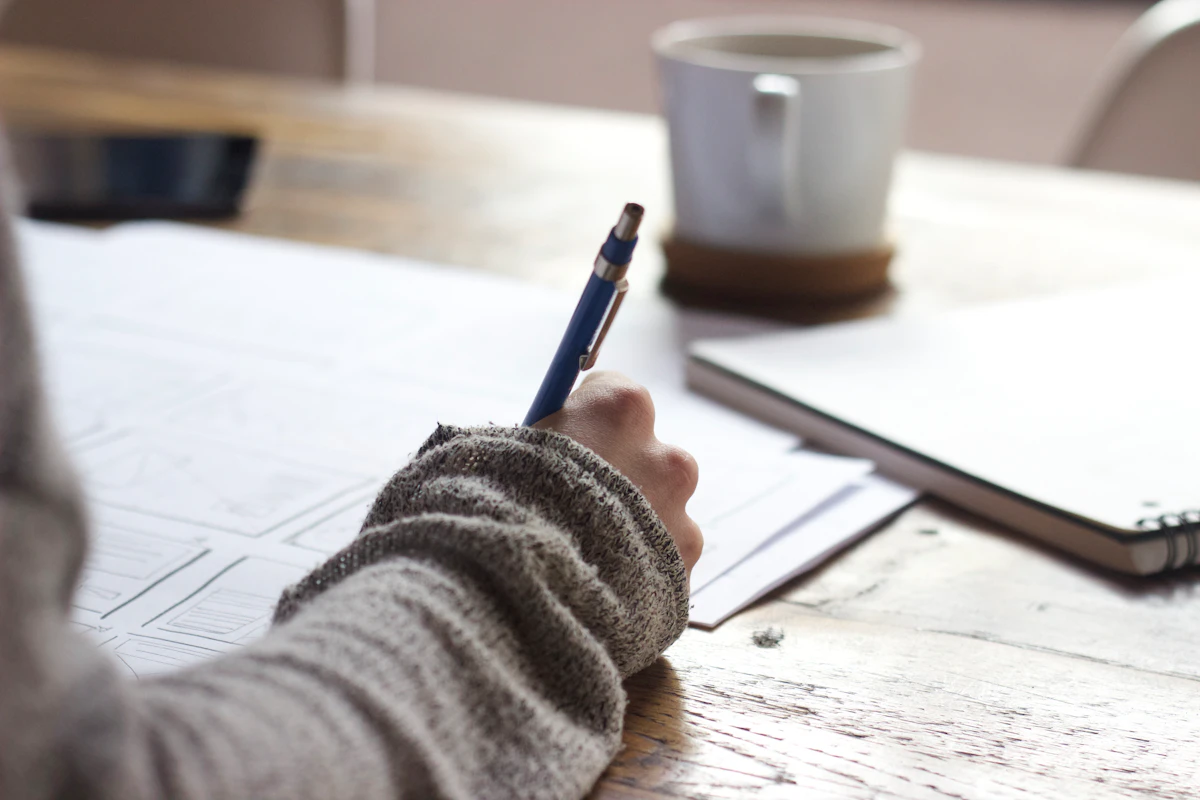 Hand in a grey sweater sketching layouts on paper beside a white mug and a notebook