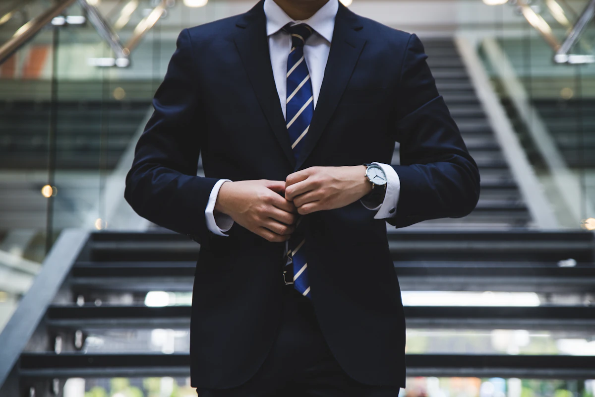 Man in a navy suit and striped tie buttoning his jacket in front of a staircase