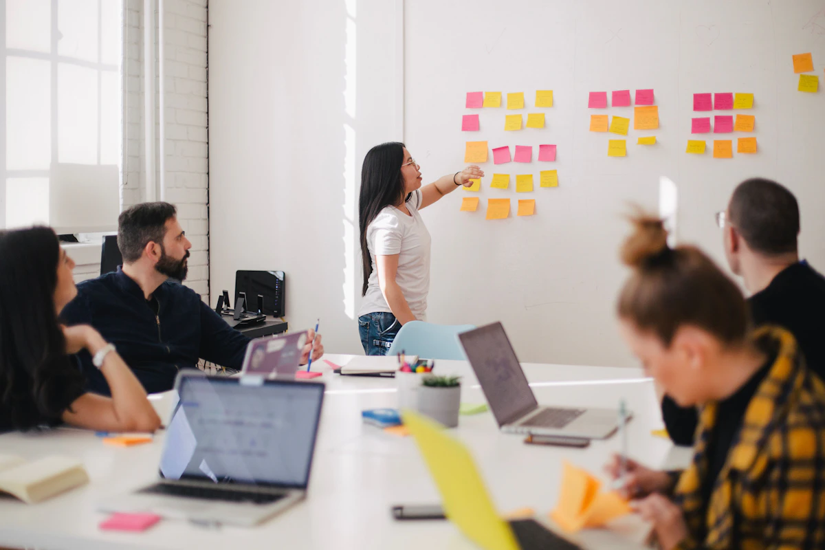 Woman pointing to sticky notes on a wall as colleagues with laptops watch from a meeting table