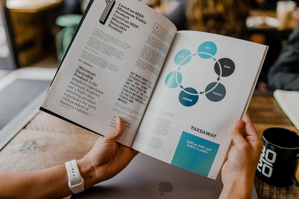 Hands holding an open magazine with a product development cycle diagram, a laptop and mug below