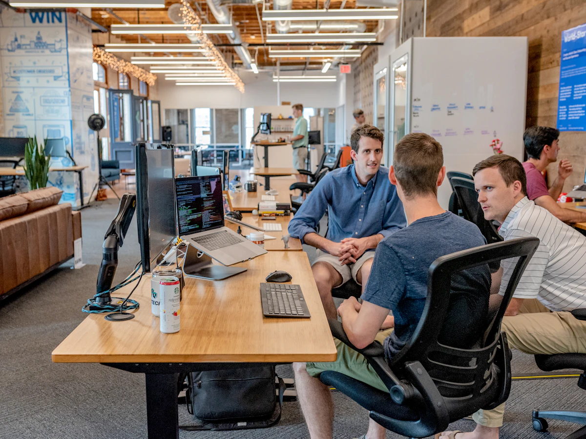 Colleagues talking at a desk with a laptop and monitors in a busy open-plan office