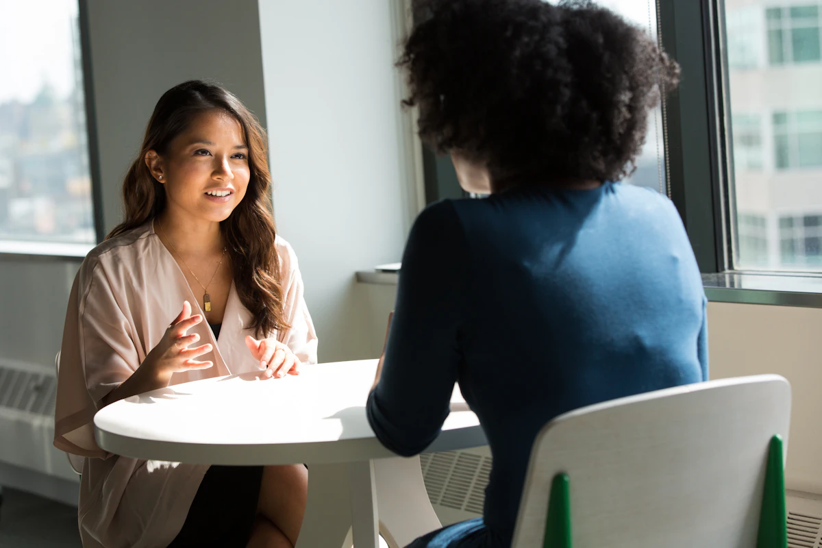 Two women talking at a small white table beside an office window